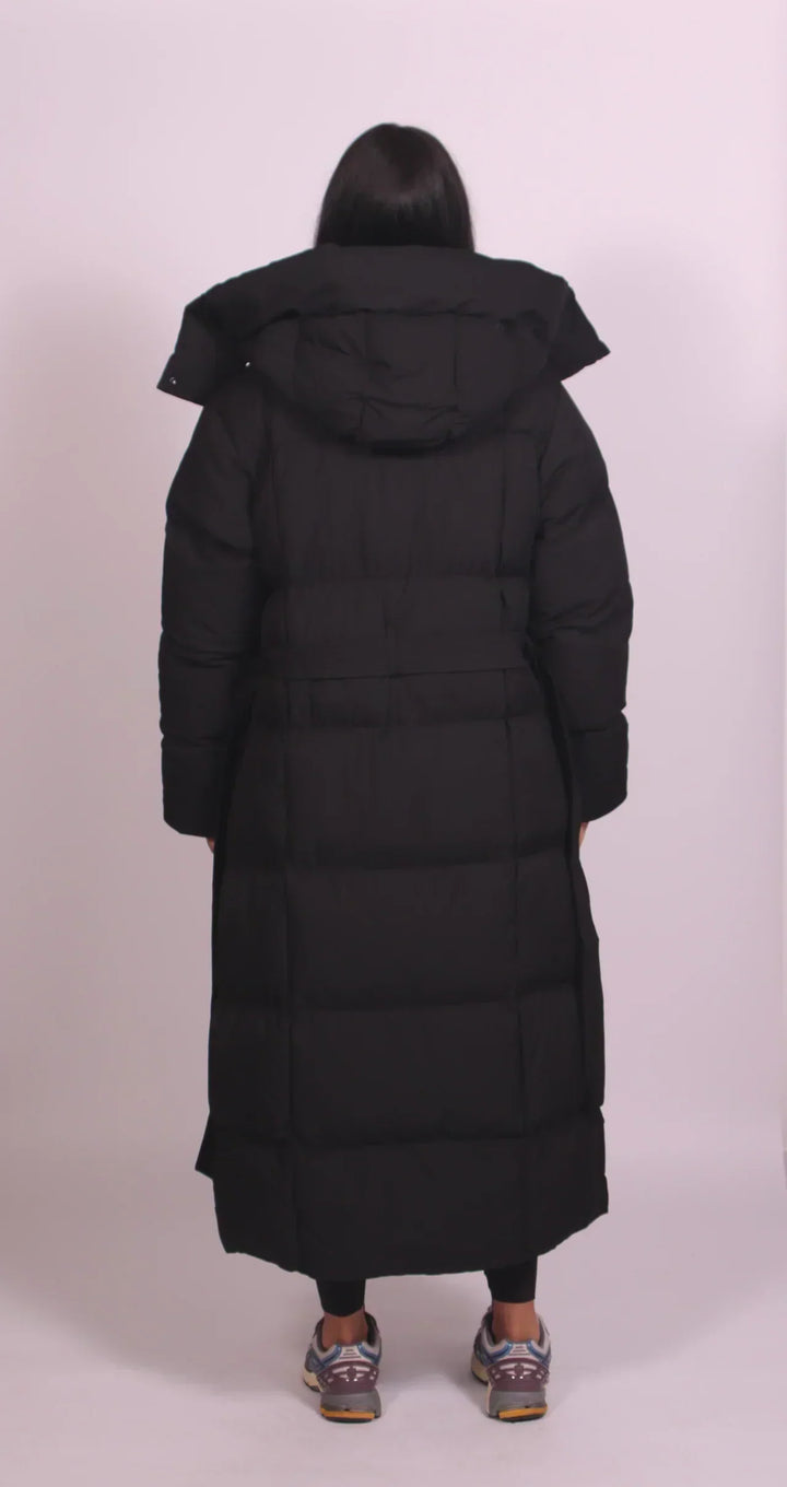Piumino lungo imbottito da donna, con cappuccio, elegante cintura e collo alto, disponibile in nero o marrone.