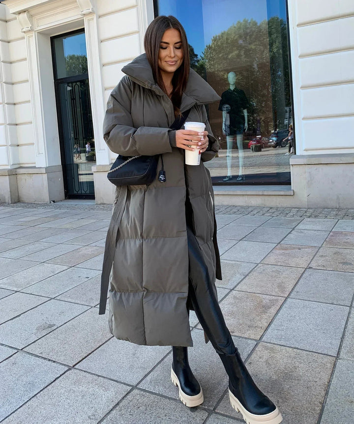 Moderno piumino lungo da donna, invernale. Collo alto e cintura regolabile per comfort e stile impeccabile.