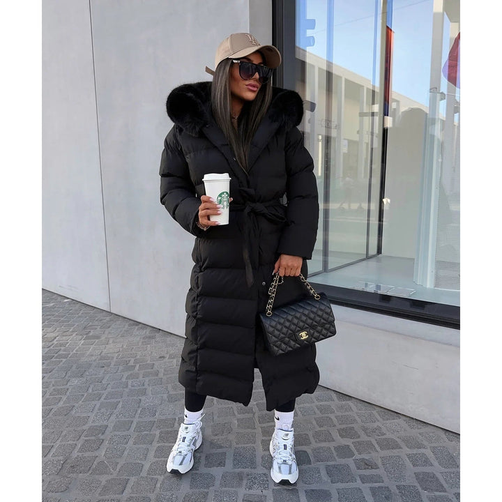 Donna indossa piumino lungo trapuntato con cintura e cappuccio in pelliccia sintetica, stile moda donna.